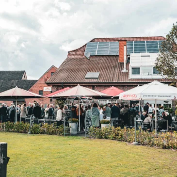 Gartenansicht mit Gästen, Bäumen und Sonnenschirmen.