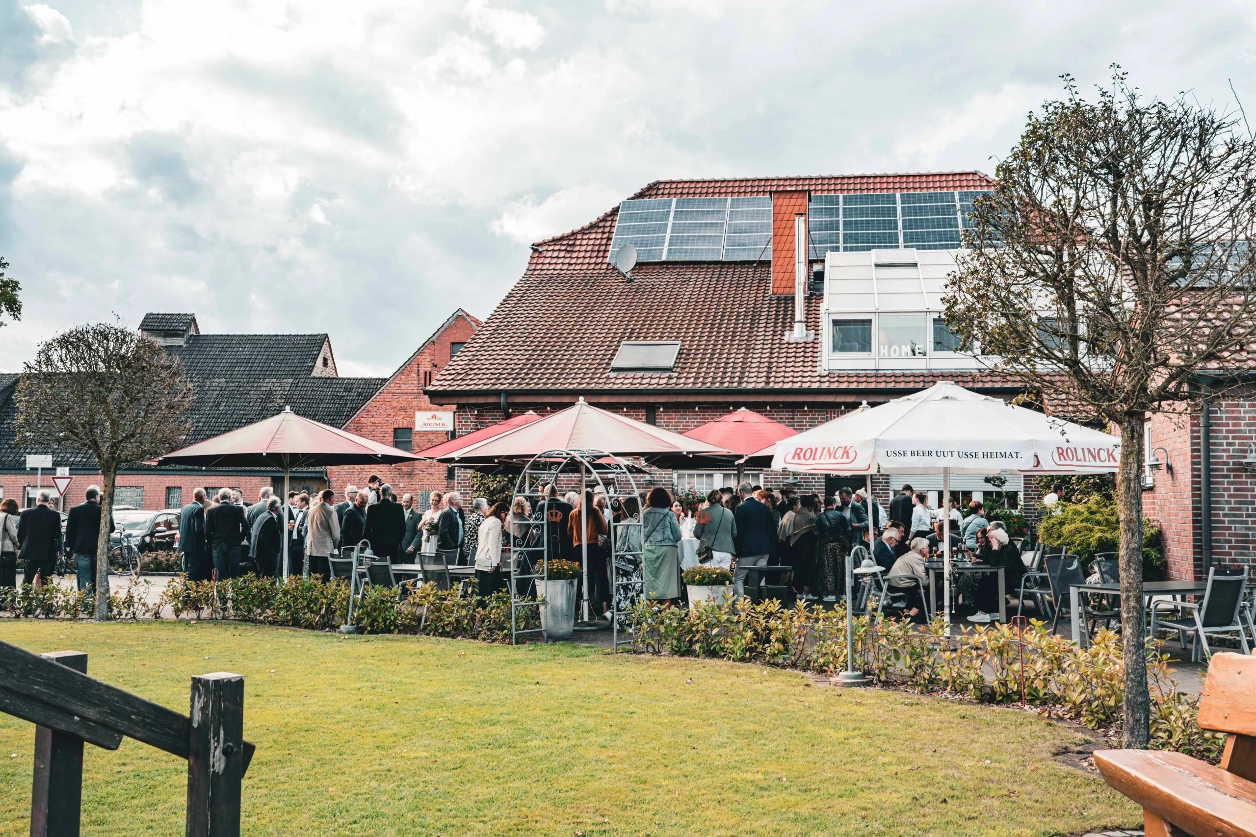 Gartenansicht mit Gästen, Bäumen und Sonnenschirmen.