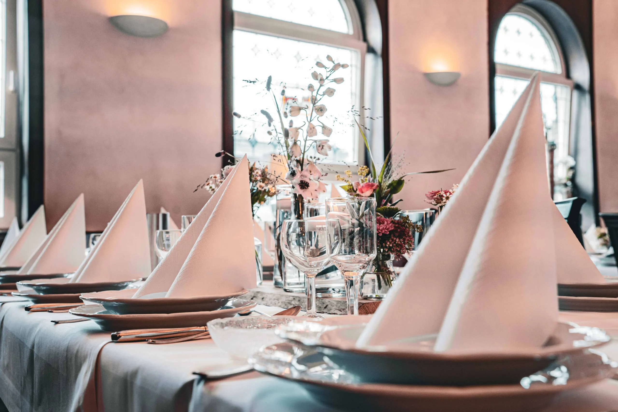 Gedeckter Tisch vor großen Bogenfenstern mit Blumenschmuck.